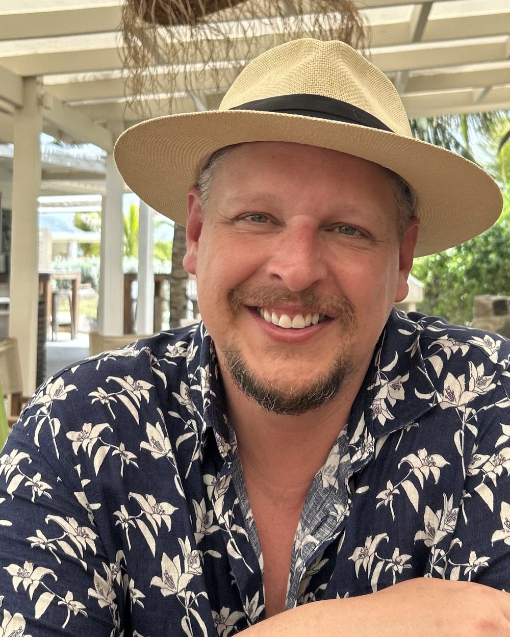 Derek Moss in a straw hat and a flowered shirt, smiling at an open-air restaurant